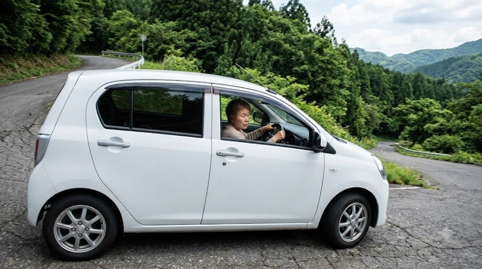 勾配のきつい山道の坂を懸命に登るダイハツ ミライースと、ハンドルを握り集中して運転する日本人高齢女性の様子。