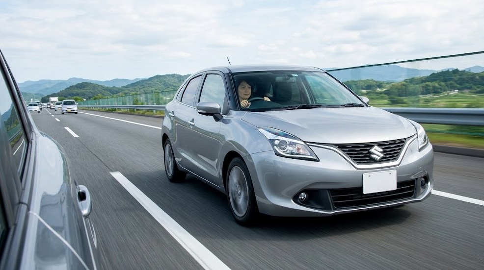 日本の高速道路を安定して走行するシルバーのスズキ バレーノ。運転席には落ち着いた表情の日本人女性。長距離ドライブの快適性と走行安定性を表現。